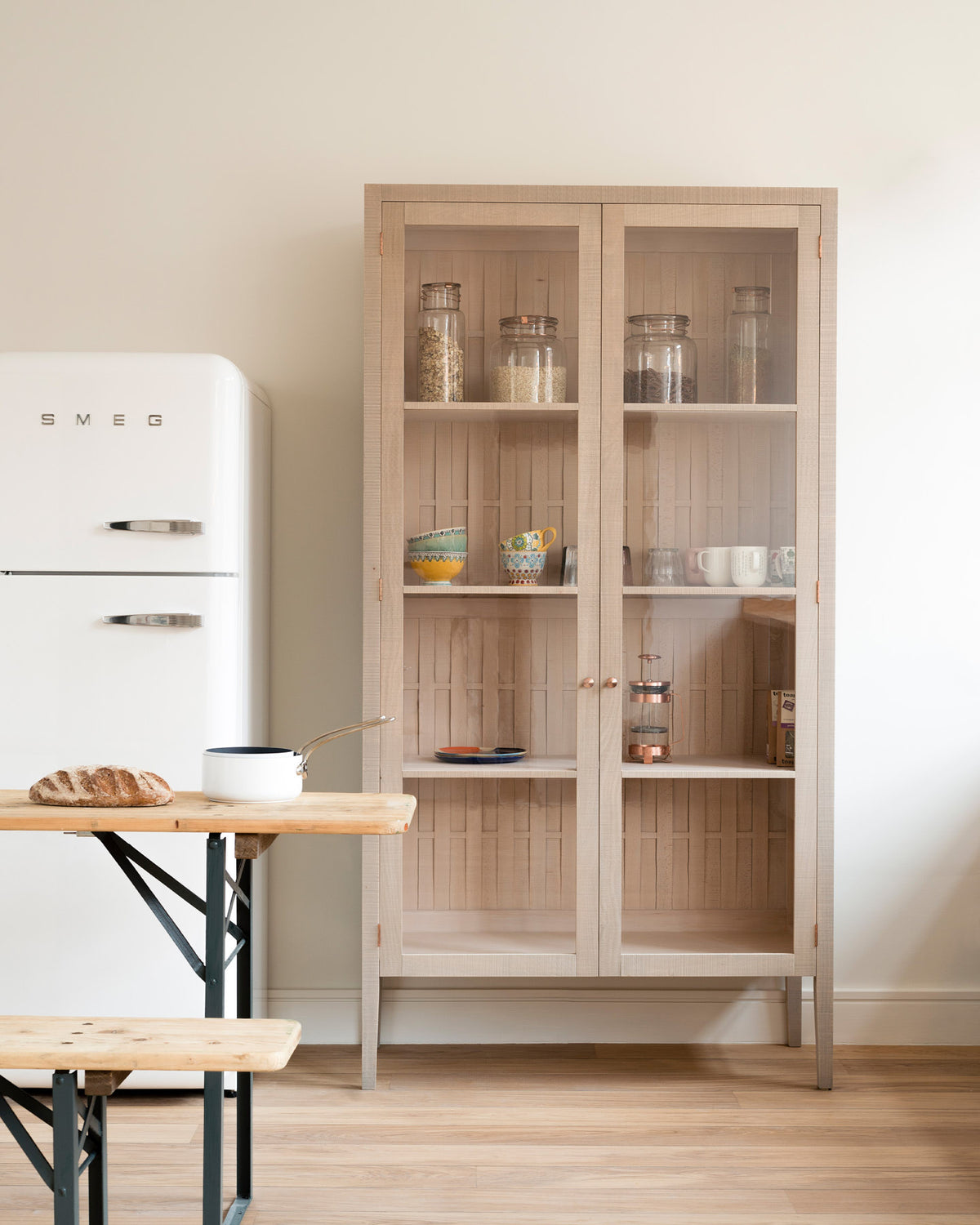 A simple and contemporary Sebastian Cox kitchen features a glazed freestanding cupboard. Adorned with colourful ceramics, a white pan, a fresh loaf of bread, large glass containers of dried ingredients, a collection of tea, a copper coffee press, and a white refrigerator, this kitchen creates a cosy space with industrial details and a practical finish.