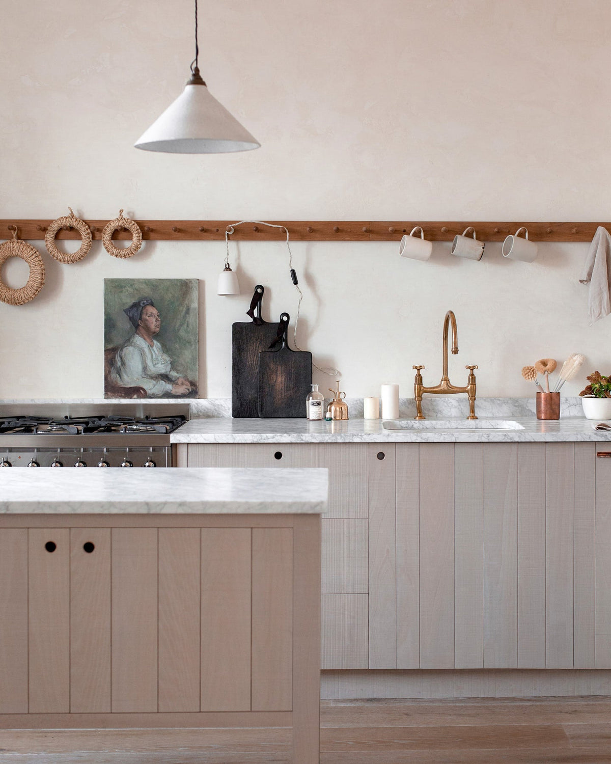 A close-up of a warm and natural Sebastian Cox kitchen features a sink and cooker run with a Carrara marble worktop, a single Belfast sink, an aged brass Ionian tap, a long oak peg rail, and a movable prep table. Beautifully decorated with artisan ceramics, a creamware rise-and-fall light, a plug-in porcelain beaker light, and blackened chopping boards, this kitchen creates a calm and delicate atmosphere that celebrates the use of natural materials.