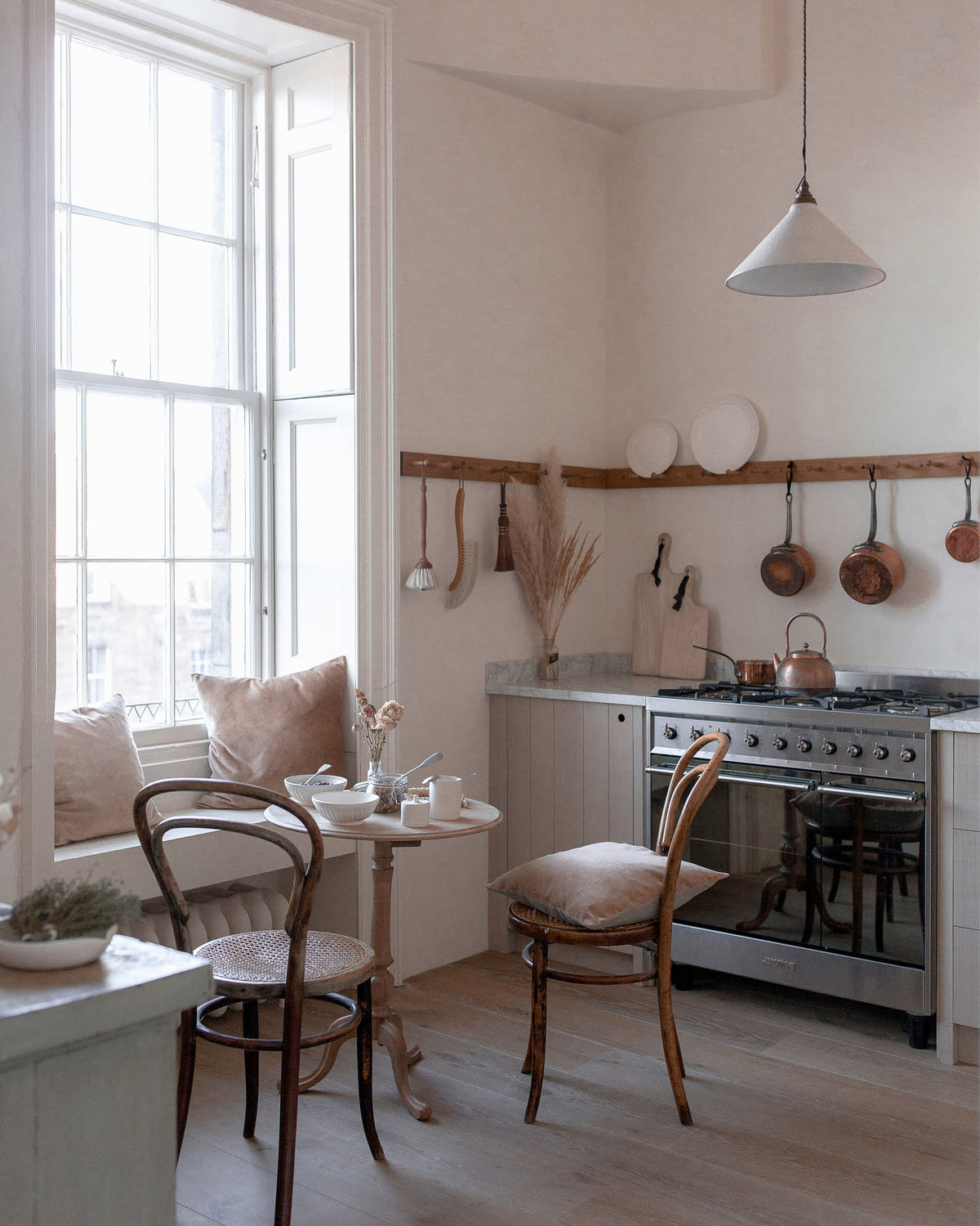 A quiet corner of a warm and natural Sebastian Cox kitchen features a sink and cooker run with a Carrara marble worktop, a single Belfast sink, an aged brass Ionian tap, and a long oak peg rail. Beautifully decorated with vintage furniture, artisan ceramics, a creamware rise-and-fall light, and pale wooden chopping boards, this kitchen creates a calm and delicate atmosphere that celebrates the use of natural materials.