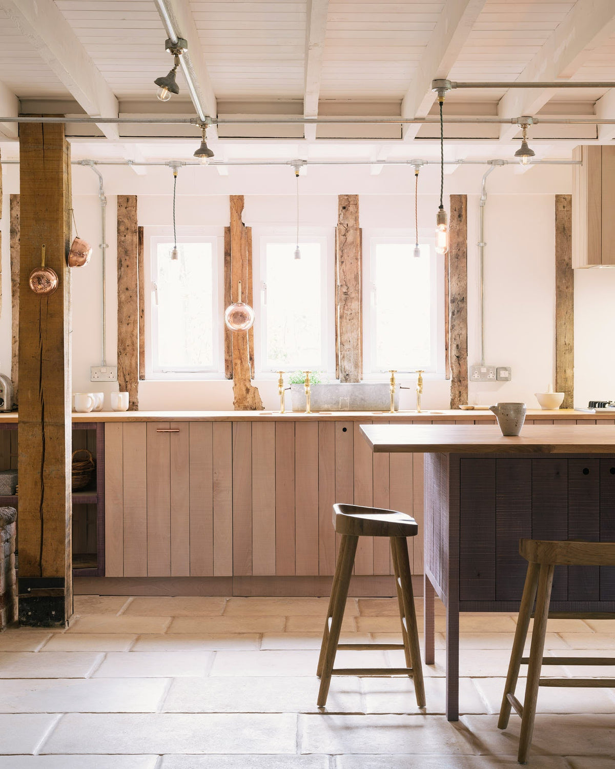 A quiet corner of a soft and spacious Sebastian Cox barn kitchen features a long sink and cooker run with a solid oak worktop, two Belfast sinks, aged brass Mayan taps, a canopy, and a kitchen island with a breakfast nook. Beautifully decorated with lavender colour tints, bare pendant lights, vintage furniture, artisan ceramics, and copper strainers, this kitchen creates an elegant and ethereal space.