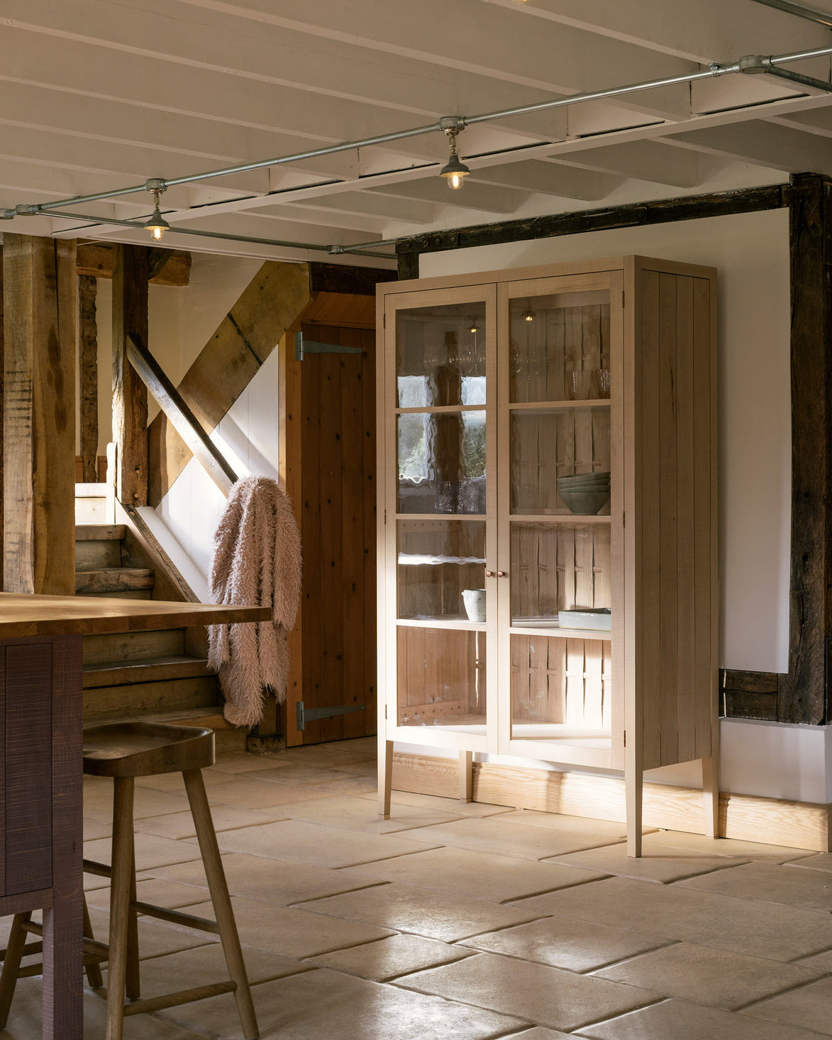 On the other side of a soft and spacious Sebastian Cox barn kitchen sits a glazed freestanding cupboard, beautifully decorated with artisan ceramics and drinking glasses. Past a small corridor is a quaint living room featuring rattan armchairs, a vintage wooden partition, and a large oak pantry cupboard.
