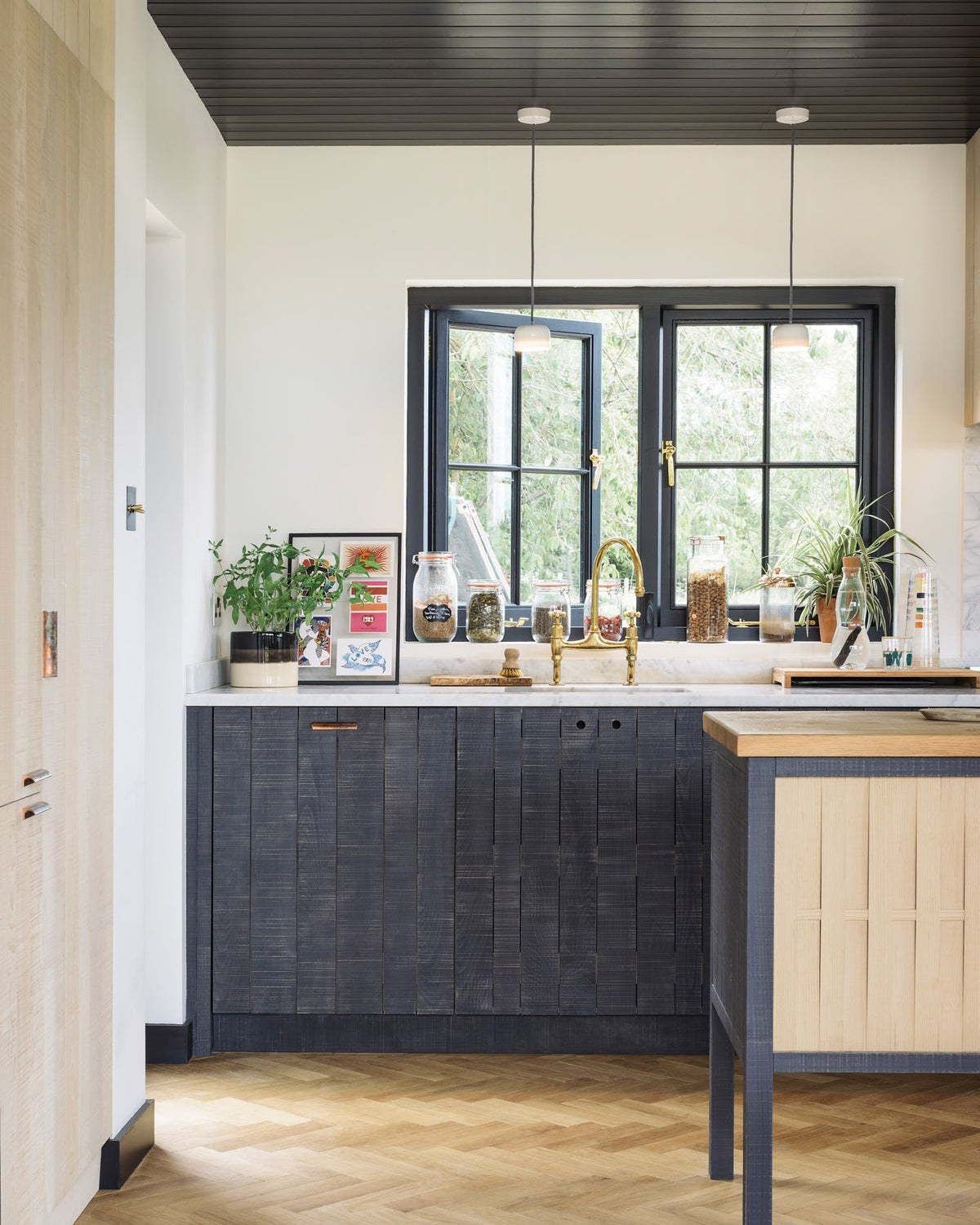 A quiet corner of a large and minimal Sebastian Cox kitchen features a long sink and cooker run with a Carrara marble worktop, a single copper sink, an aged brass Ionian tap, a canopy, a built-in bank of cupboards, and a large kitchen island with a solid oak worktop. Beautifully decorated with artisan ceramics, dried ingredients in glass jars, a water carafe, and drinking glasses, this kitchen creates a simple and practical space.