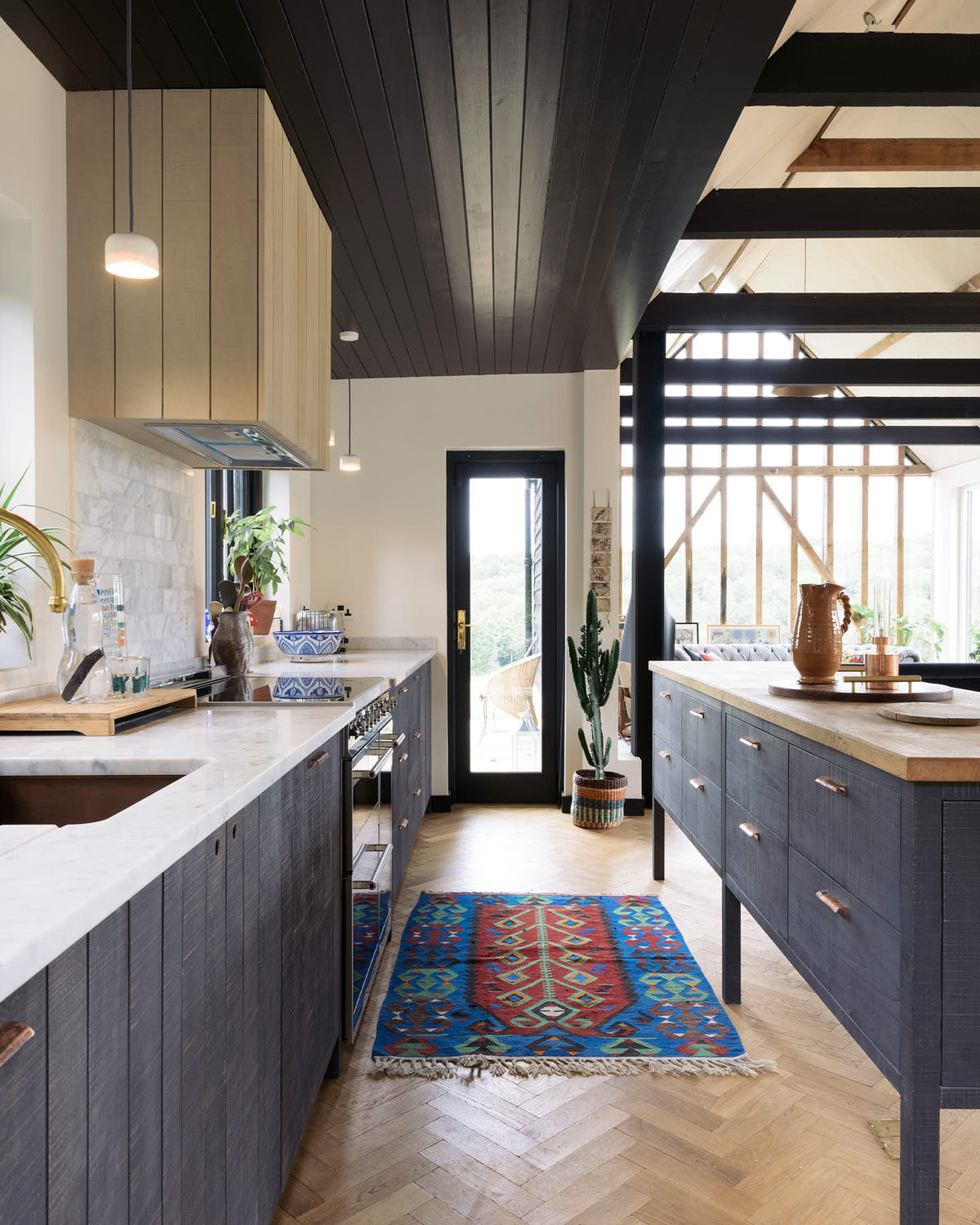 Nestled between a large and minimal Sebastian Cox kitchen, which features a long sink and cooker run with a Carrara marble worktop and a large kitchen island with a solid oak worktop, is a slim glass door leading to the gorgeous countryside of Tuscany. Beautifully decorated with artisan ceramics, a large cactus plant, a wicker basket, and a bright patterned rug, this kitchen creates a simple and practical space.
