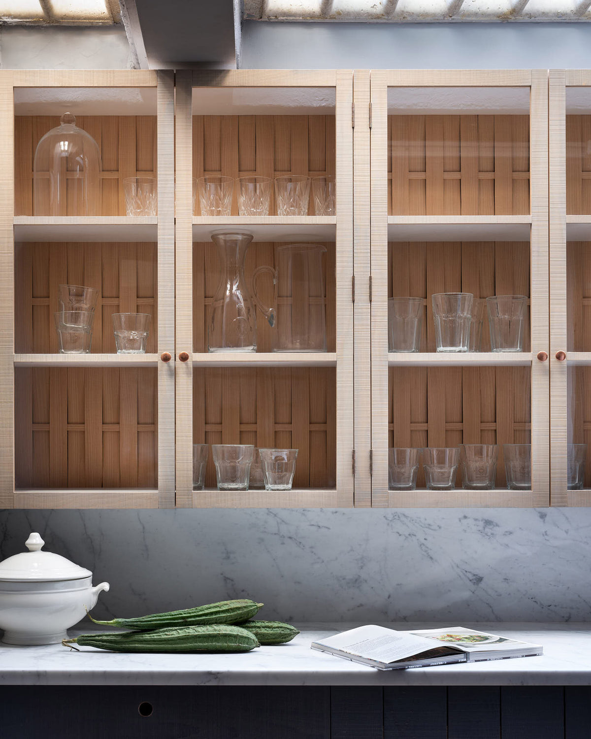 A closer look at a chic and simple Sebastian Cox basement features a long sink run with Carrara marble countertop and splashback, and glazed wall cupboards. This kitchen creates a cool and atmospheric space, beautifully decorated with artisan ceramics, drinking glasses, a book, and bitter melons.
