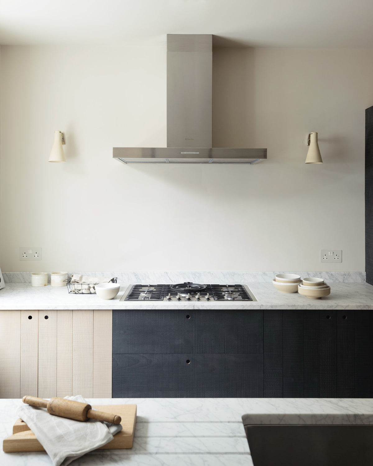 Past a black-framed transom partition window is a sleek and modern Sebastian Cox galley kitchen featuring a sink and cooker run with a Carrara marble worktop. Adorned with simple ceramics, beaker wall lights, a metal canopy, a chopping board, a rolling pin, and tea towels, this kitchen creates an elegant and practical space.
