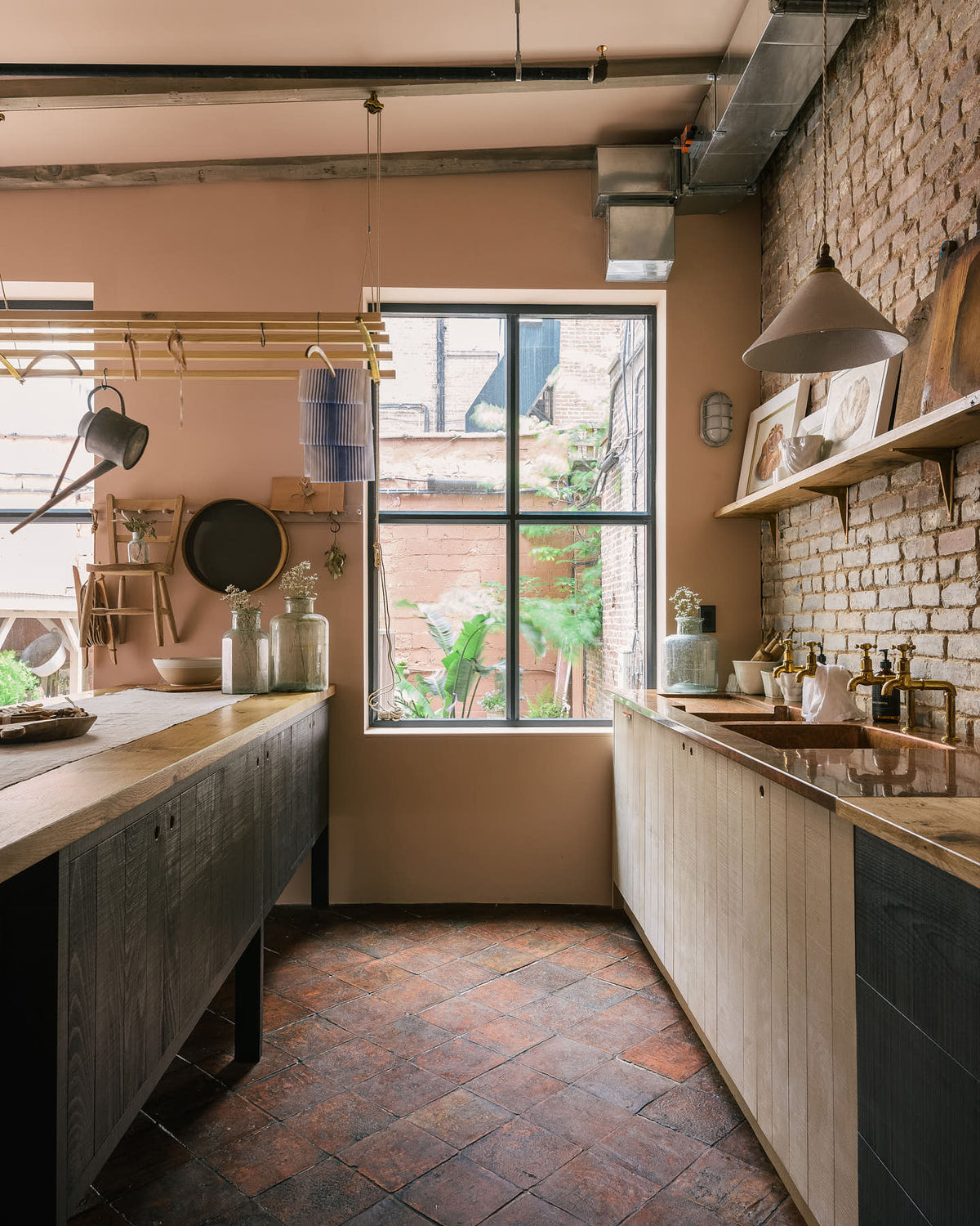 Nestled between the long sink run of a calm and simple Sebastian Cox kitchen, which features two sets of copper sinks and aged brass Mayan taps, a copper worktop, and a prep table with a solid oak worktop, this kitchen is adorned with vintage items, artisan ceramics, wooden chopping boards, framed illustrations, and a terracotta square floor. It creates a chic New York industrial space that celebrates a love of natural materials.