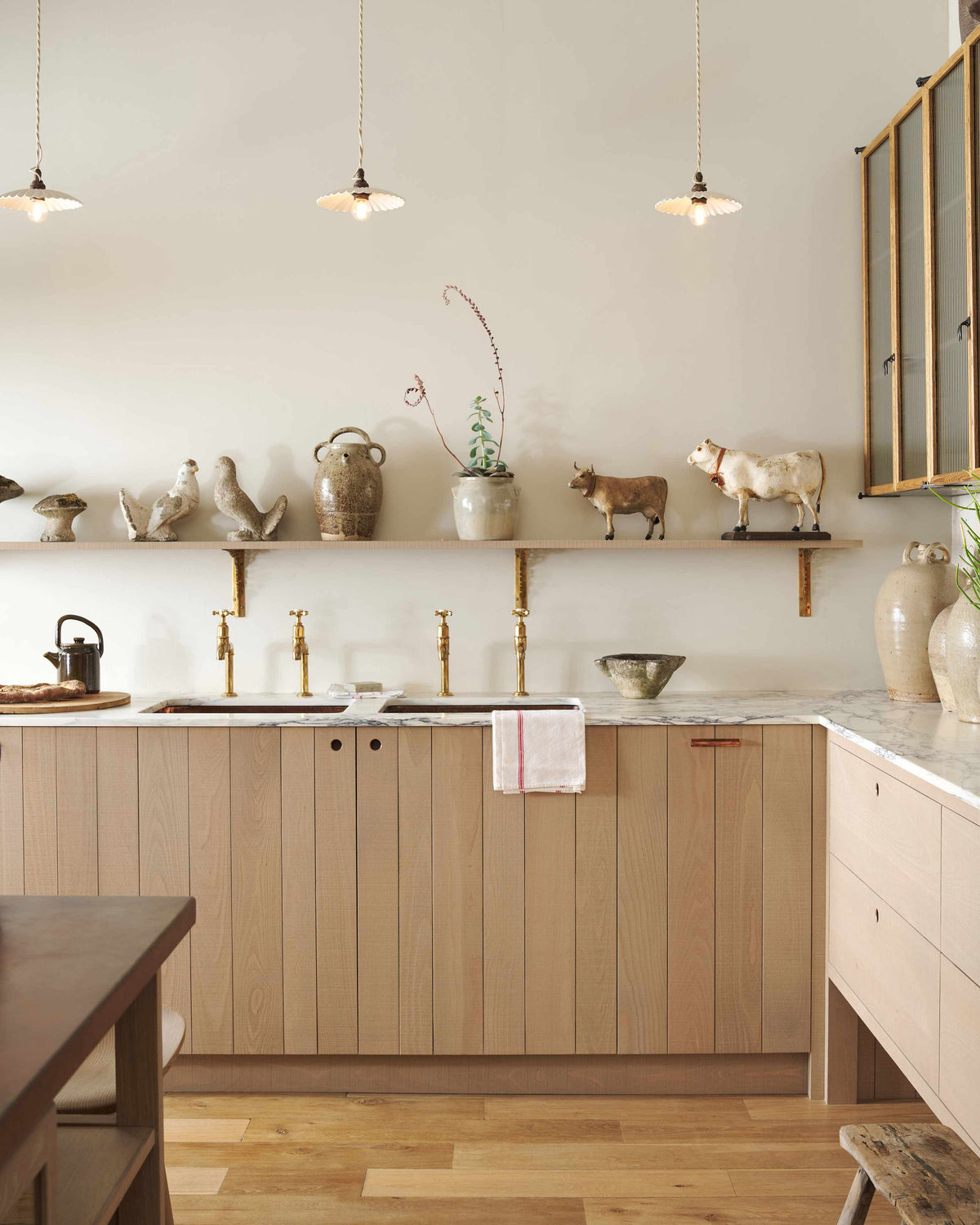 An all-natural and functional Sebastian Cox kitchen features a cooker and sink run with a marble worktop, glazed wall cupboards, a free-standing pantry with glazed doors and an iconic Haberdasher’s prep table with a copper worktop. Lovingly adorned with antiques, artisan ceramics, woven baskets, and beautiful houseplants, it creates a wonderfully simple kitchen with a love of natural sustainability.