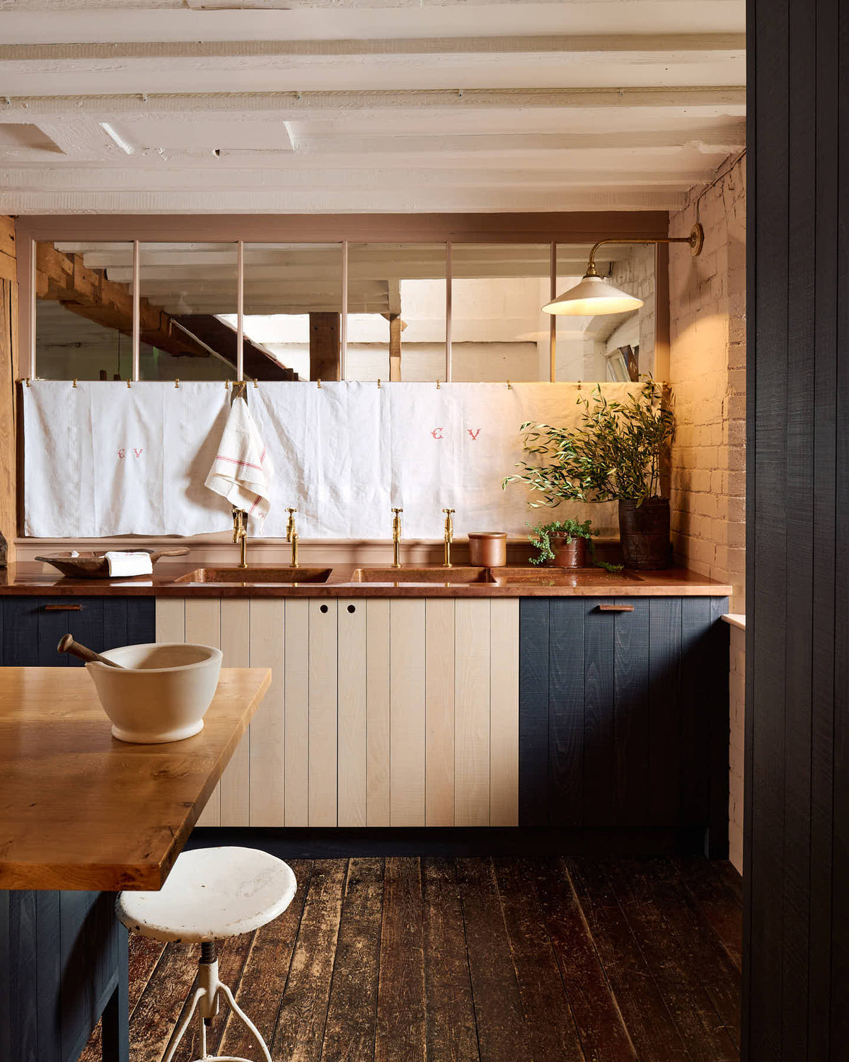 A warm and stylish Sebastian Cox kitchen features a sink run with two single copper sinks, aged brass Mayan taps, glazed partitions, a copper worktop, and a preptable with a solid oak worktop. Adorned with an aged brass café curtain rail, a milk glass task light, artisan ceramics, and vintage wooden items, this kitchen creates a humble space that celebrates a love of simple and natural materials.