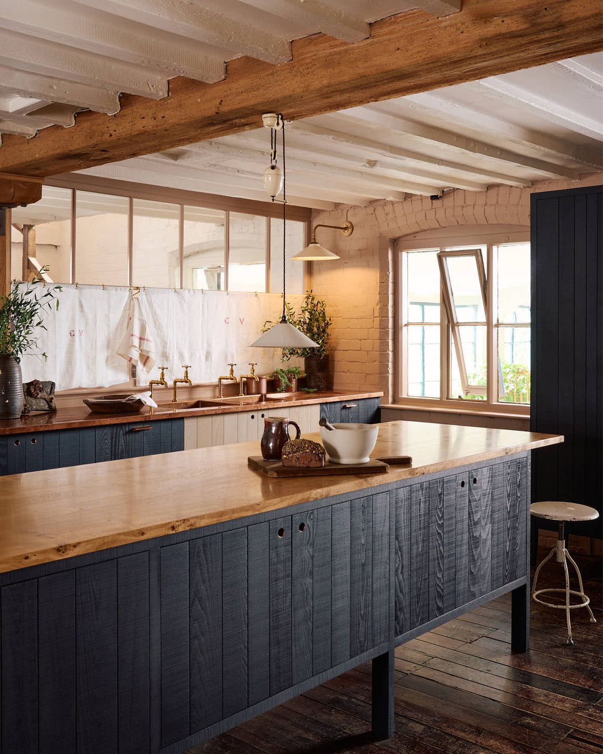 An oblique view of a warm and stylish Sebastian Cox kitchen features a sink run with two single copper sinks, aged brass Mayan taps, glazed partitions, and a copper worktop. Adorned with an aged brass café curtain rail, a milk glass task light, a creamware rise and fall light, artisan ceramics, and vintage wooden items, this kitchen creates a humble space that celebrates a love of simple and natural materials.