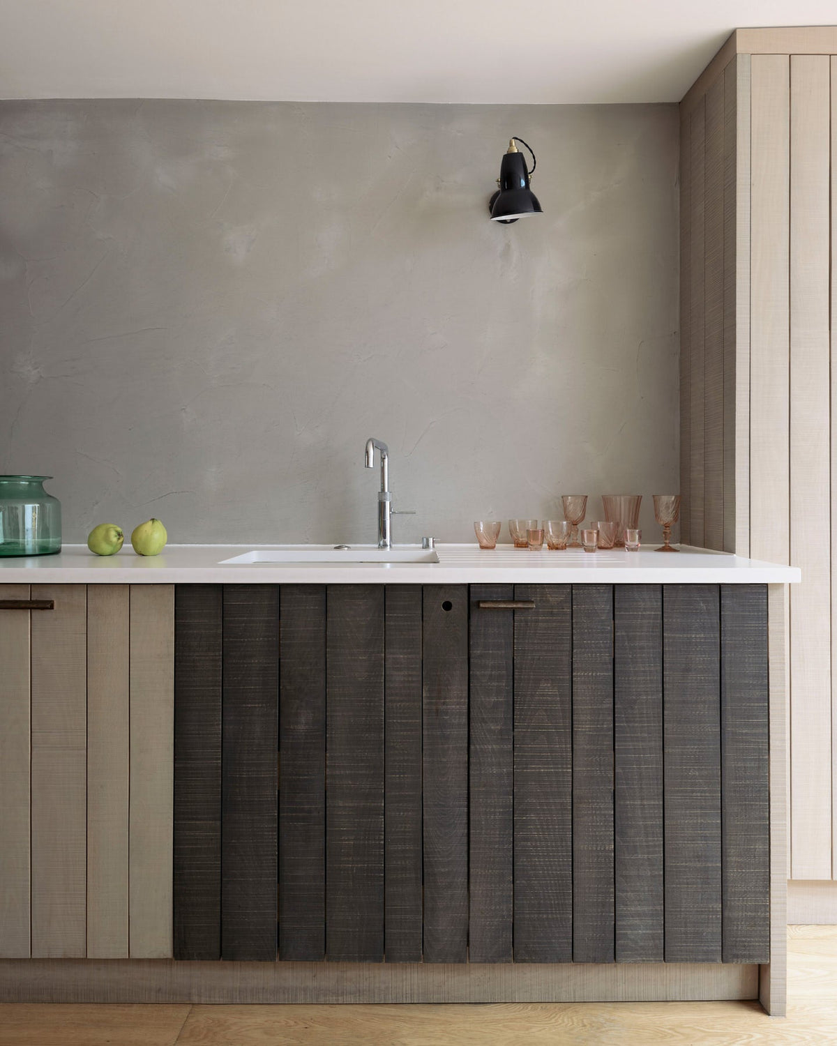 A close-up of a sleek and simple Sebastian Cox kitchen includes a cooker and sink run topped with a silestone worktop, a full-height larder, a single Belfast sink, a silver tap, and walls finished with natural limestone. The space is beautifully decorated with red-tinted drinking glasses and a black industrial wall lamp, creating a minimal yet earth-conscious and sustainable kitchen environment.