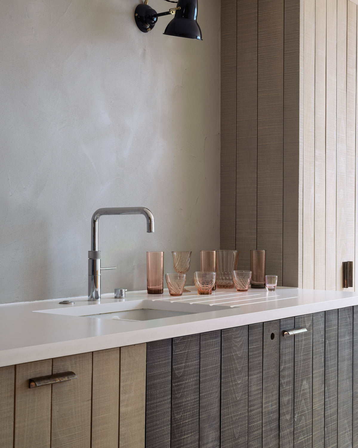 An oblique view of a sleek and simple Sebastian Cox kitchen includes a cooker and sink run topped with a Silestone worktop, a full-height larder, a single Belfast sink, a silver tap, and walls finished with natural limestone. The space is beautifully decorated with a plate of fresh green pears, red-tinted drinking glasses, copper shot glasses, a large blue vase, and a copper bowl, creating a minimal yet earth-conscious and sustainable kitchen environment.