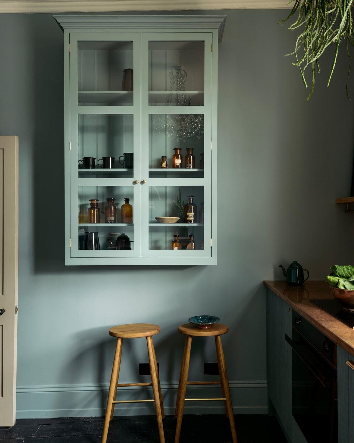 A sweet and simple Clerkenwell blue Sebastian Cox kitchen features a short cooker and sink run with a copper worktop and splashback, and a glazed wall cupboard. Adorned with artisan ceramics, vintage bric-a-brac, hanging plants, a framed quirky illustration, and Helen stools, this kitchen creates a glamorous and practical space.