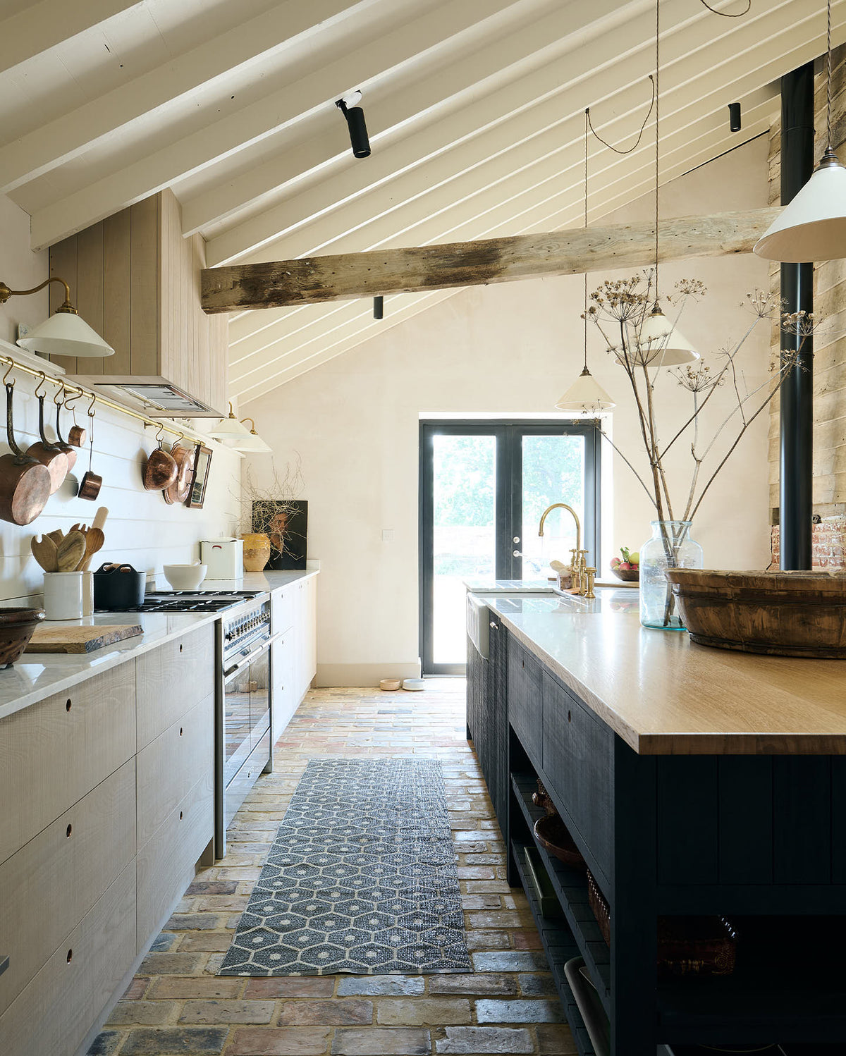 A beautifully renovated farm features a Sebastian Cox kitchen with a long cooker run, a large kitchen island with a double fluted Belfast sink, an aged brass Ionian tap, and a Carrara marble and solid oak worktop. Adorned with creamware pendant lights, vintage items, artisan ceramics, and a bowl of apples, this kitchen creates a clean and contemporary style that harmonises with the natural materials used throughout the space.