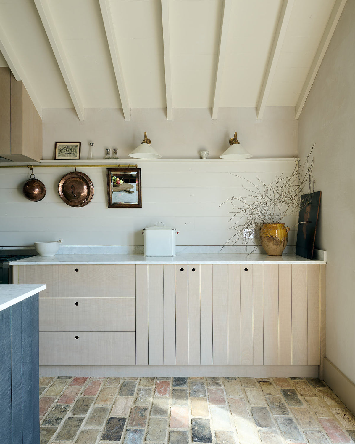 A quiet corner of a beautifully renovated farm features a Sebastian Cox kitchen with a long cooker run, a large kitchen island with a double fluted Belfast sink, an aged brass Ionian tap, and a Carrara marble and solid oak worktop. Adorned with Heirloom wall lights, vintage items, artisan ceramics, and an oil painting, this kitchen creates a clean and contemporary style that harmonises with the natural materials used throughout the space.