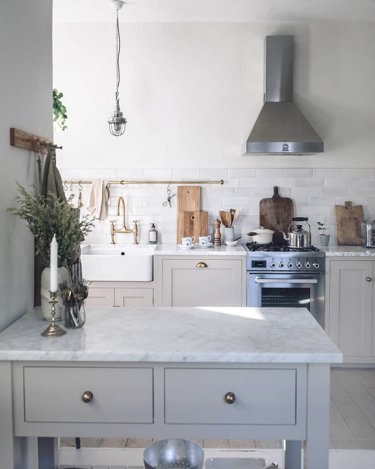A humble Mushroom colour shaker kitchen with an all-in-one L-shaped sink and cooker run, and a small rolling kitchen island with Carrara marble worktop. A single Belfast sink is complemented by an aged brass Ionian tap, and an aged brass hanging rail adds a striking element against the grey and neutral tones of this charming kitchen.