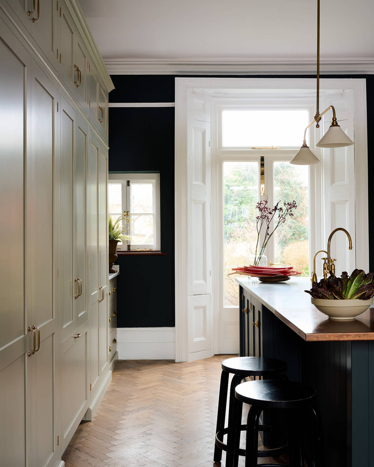 A quiet corner of a simple and elegant mushroom-brown Shaker kitchen features a cooker run with freestanding cupboards, a Carrara marble worktop, a false chimney breast, white porcelain splashback tiles, and a large kitchen island. The space feels loving and functional, adorned with artisan ceramics, copper pans, bright red rhubarb, luscious houseplants, and books.