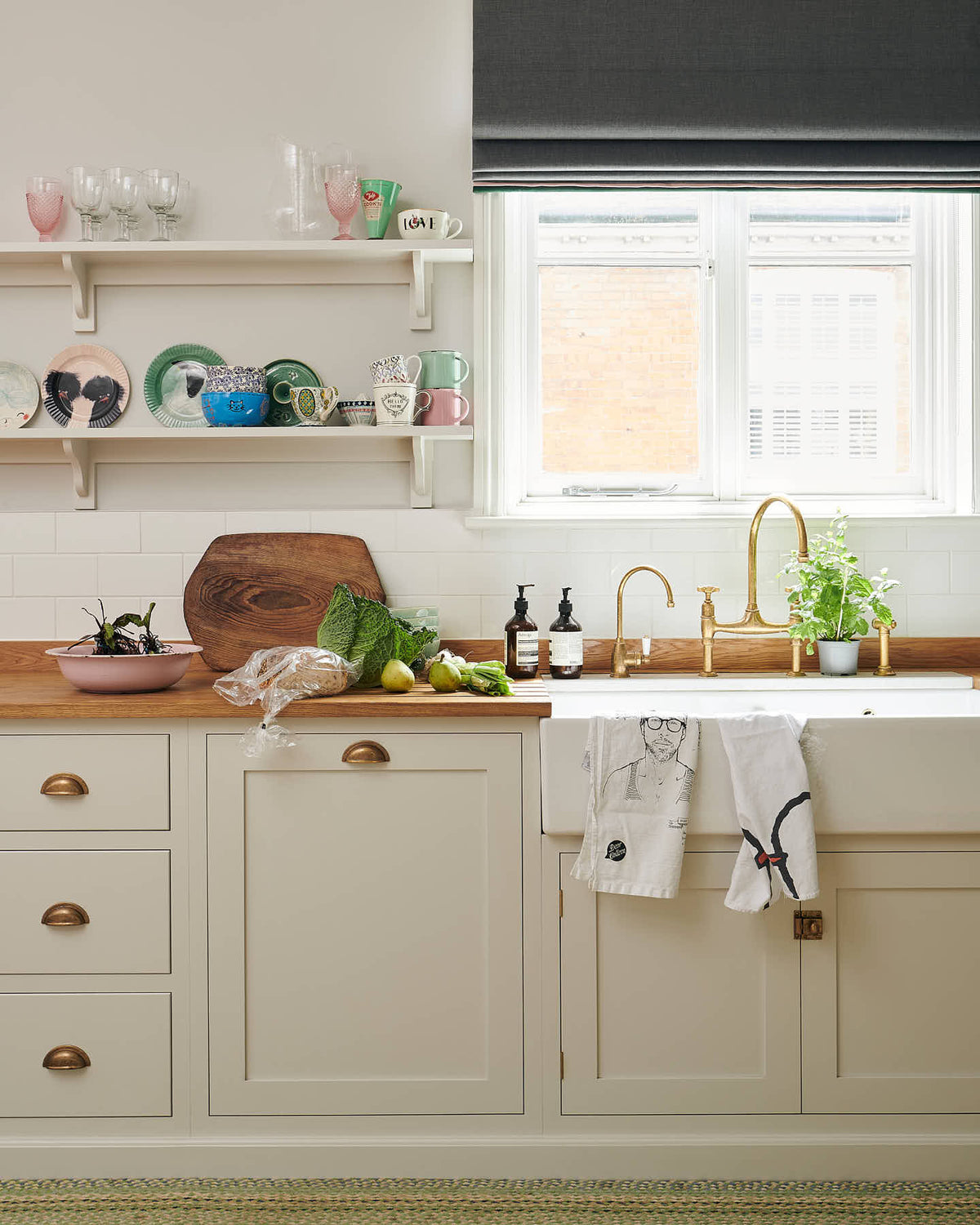 A vibrant Shaker kitchen features an L-shaped sink and cooker run with a solid oak worktop, a single Belfast sink, an aged brass Ionian tap, and Shaker shelves. Lovingly decorated with beautiful ceramics and drinking glasses, luscious green vegetables, and illustrative tea towels, it creates a characterful and inviting space.