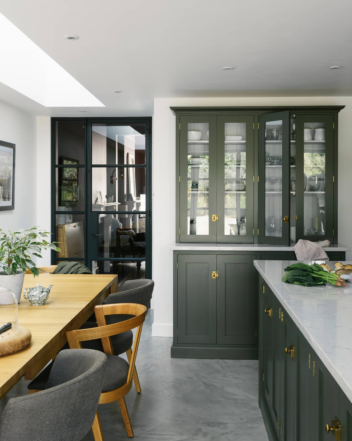 A bright and sophisticated Bakehouse Green shaker kitchen, styled with Scandinavian furniture and a soothing green palette. The kitchen features a central island with a double Belfast sink and a glazed larder with Silestone worktop. There’s a beautiful skylight above the solid oak dining table.