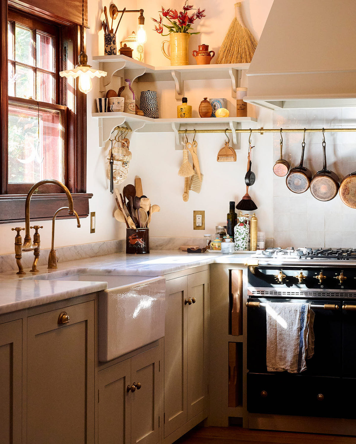 A tight and busy corner of a humble mushroom-painted shaker kitchen between the L-shaped cooker and sink run with a Carrara marble worktop. The shaker shelves with aged brass hanging rails hold all sorts of kitchen items, from a triangle bell and a small basket of garlic to measuring spoons, cleaning brushes, and pans.