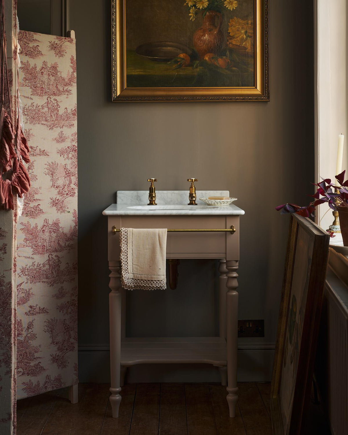 An oblique view towards a dusty-pink Victorian-inspired washstand with a brass mixer tap in a greyish-brown painted room. The shot is atmospheric and moody, with two lit candles casting a subtle glow and light from an adjacent window illuminating the brass accents. | Pillar Taps shown TEST