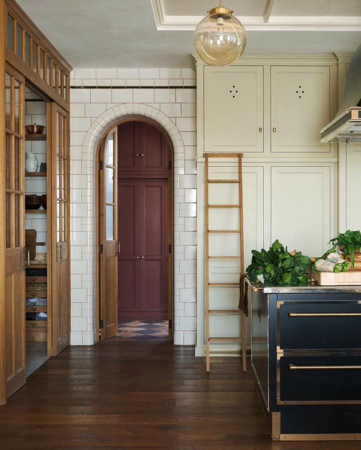 Tall fitted Classic cupboards, painted in a creamy stain colour, with an elegant oak ladder sit next to a curved archway tiled with glossy white tiles in a traditional brickbond pattern. A tall glazed oak door is open into the next room to reveal tall burgundy painted Classic cupboards and a moody chequerboard marble floor.