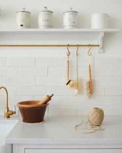 An assortment of kitchen utensils hang from a brass rail in front of white metro wall tiles with delicate crazing.