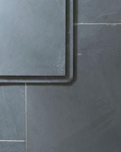 A close-up overhead view of a grey tumbled slate floor, with two loose tiles stacked in one corner.