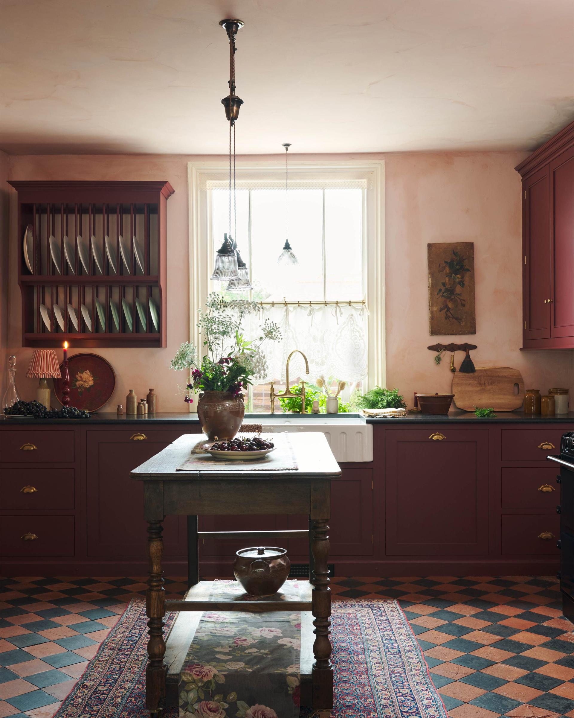 A warm and simply gorgeous refectory-red Shaker kitchen features a sink run with a black granite worktop, a wall cupboard, a small Heirloom plate rack, a pantry cupboard, and a gas stove in a chimney breast with Sepia Lace market splashback tiles. Beautifully decorated with vintage furniture, artisan ceramics, original botanical illustrations, reeded glass pendant lights, and a wild floral arrangement, it creates a humble and endearing space.