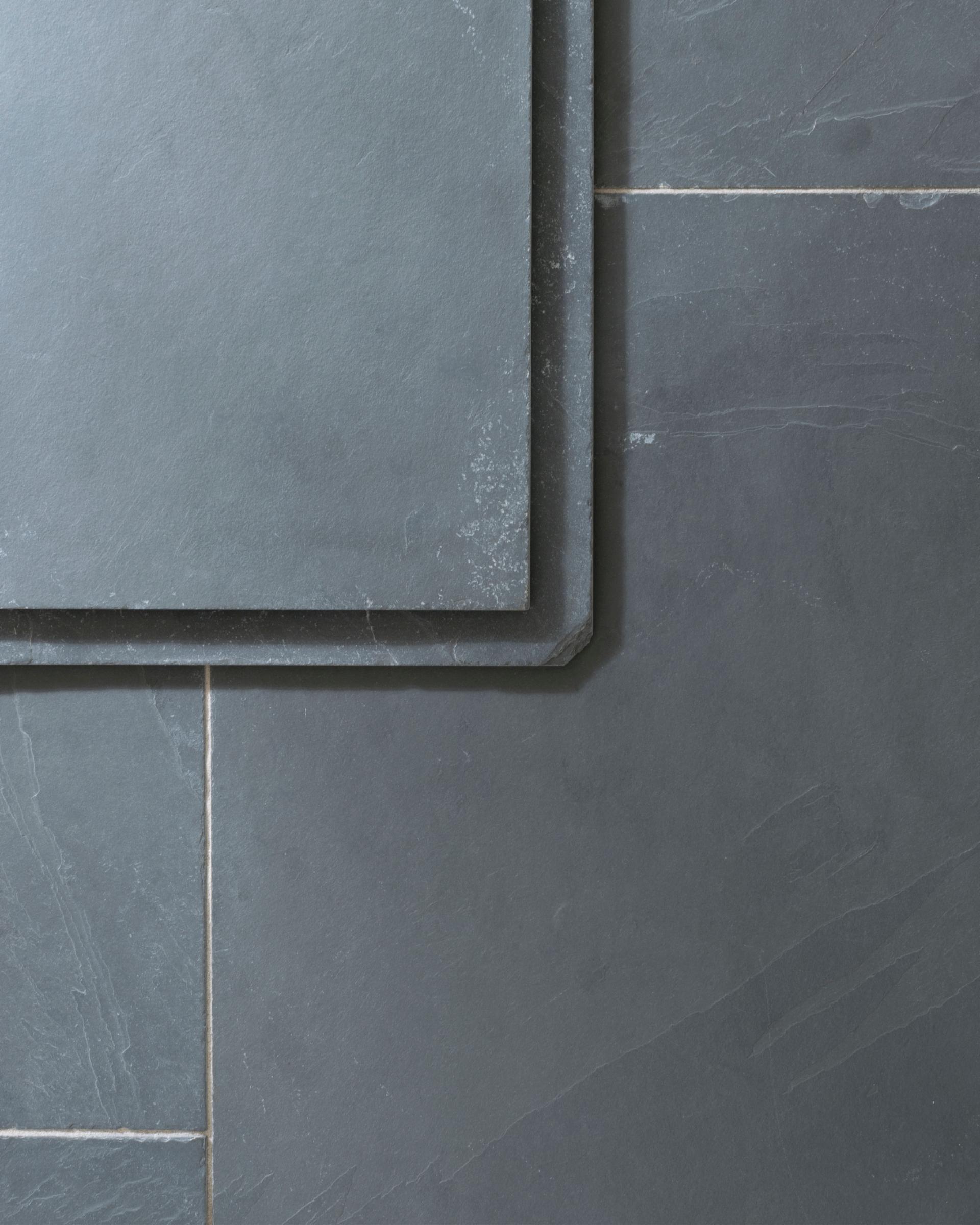A close-up overhead view of a grey tumbled slate floor, with two loose tiles stacked in one corner.