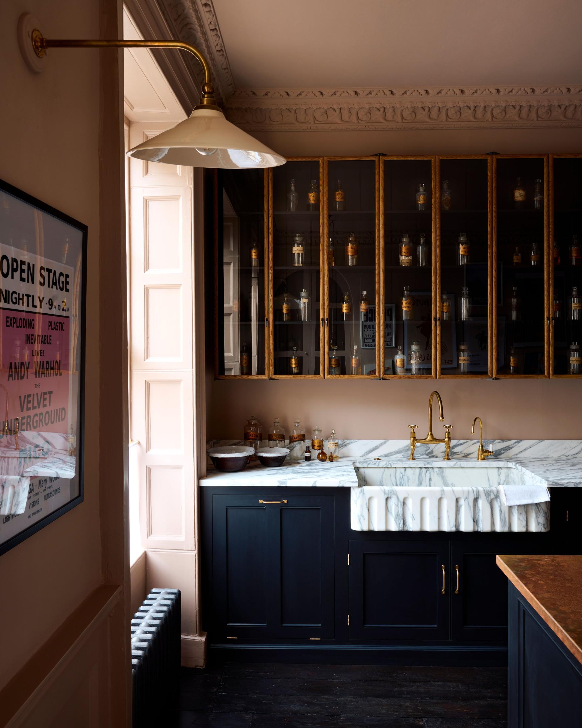 A straight-on view of a black painted Georgian-style kitchen in an atmospheric room with dark floorboards, pink walls and tall mid-century-style glazed oak wall cupboards.