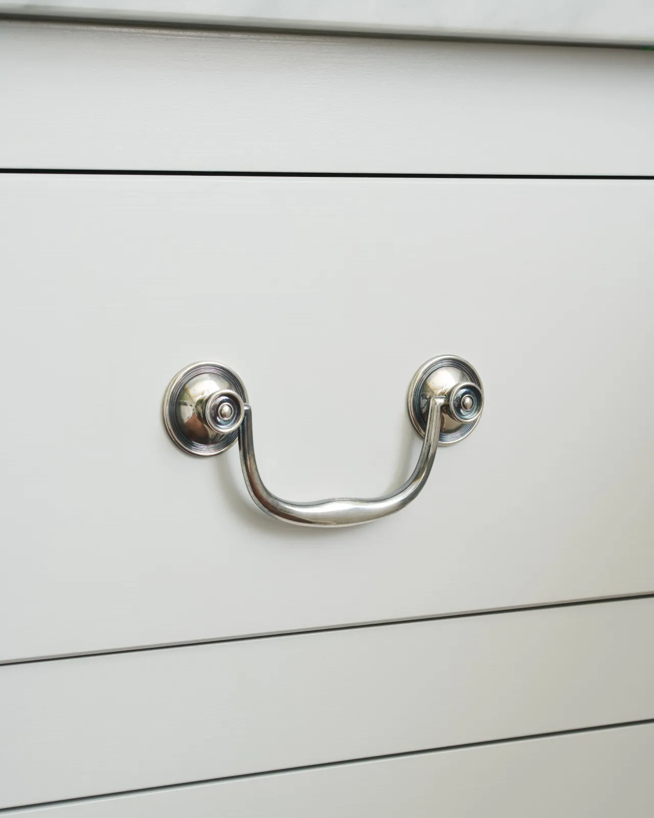 An angled view of an elegant Georgian-style silver swing handle fitted to a white painted kitchen drawer.