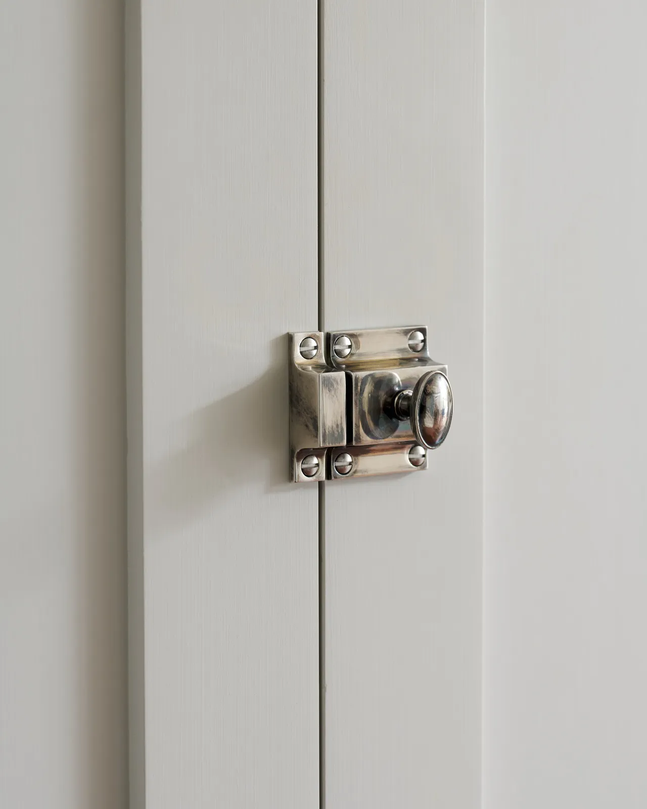 A left side-on view of a classic cupboard catch in a tarnished silver finish, fitted to a glazed white cupboard.