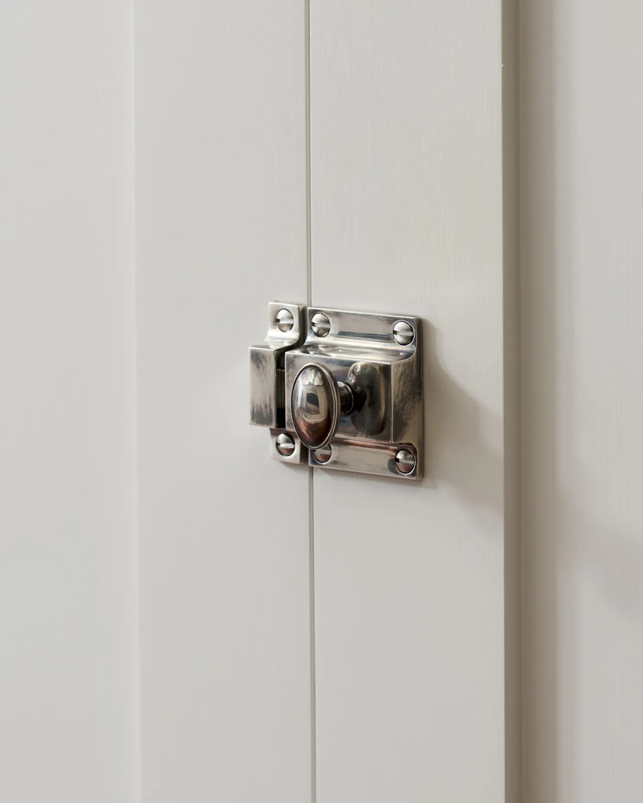 A right side-on view of a classic cupboard catch in a tarnished silver finish, fitted to a glazed white cupboard.