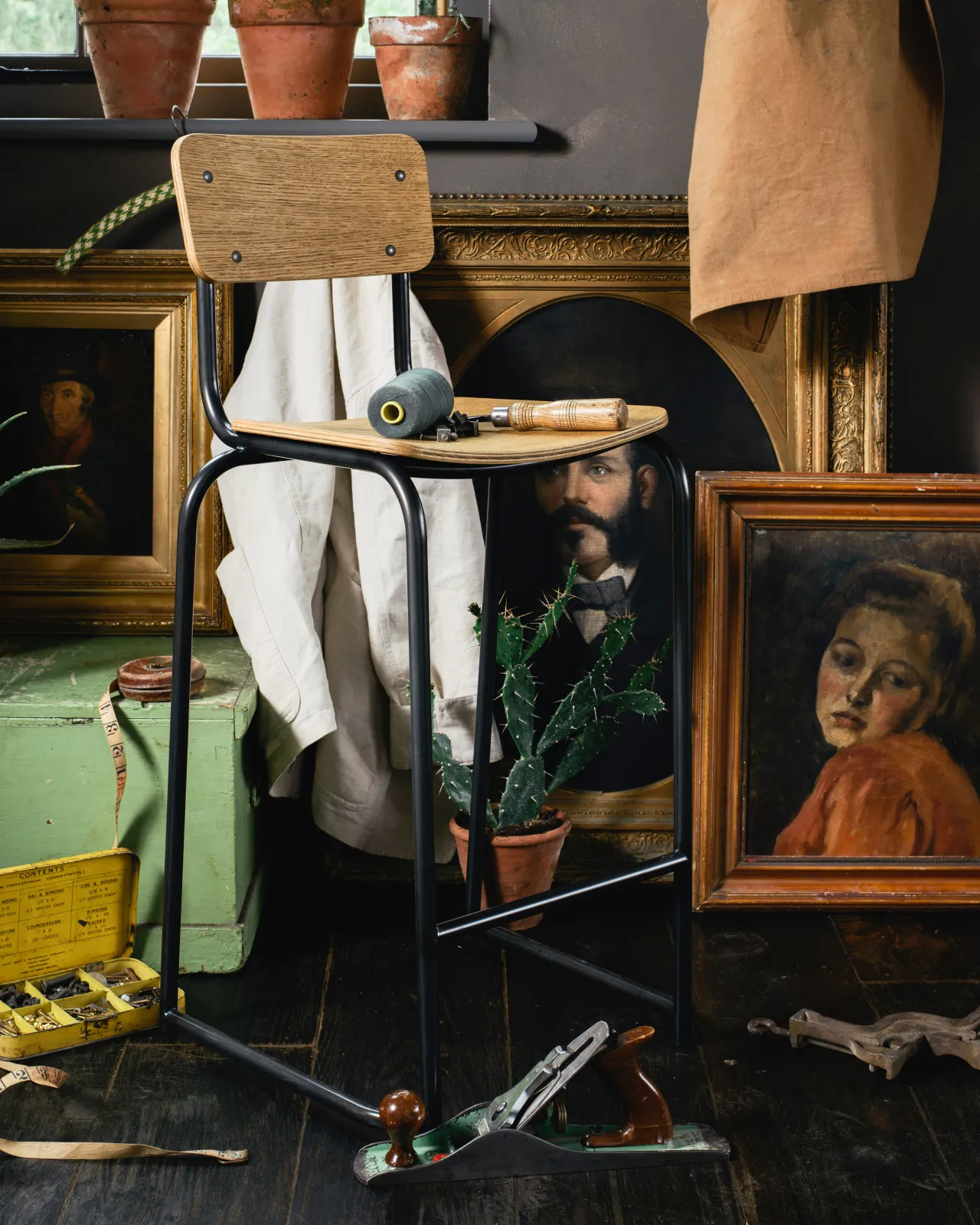 A mid-century-style industrial bar stool with a black tubular metal frame and shaped oak veneer plywood seat and backrest in a dark room with antique paintings and furniture.