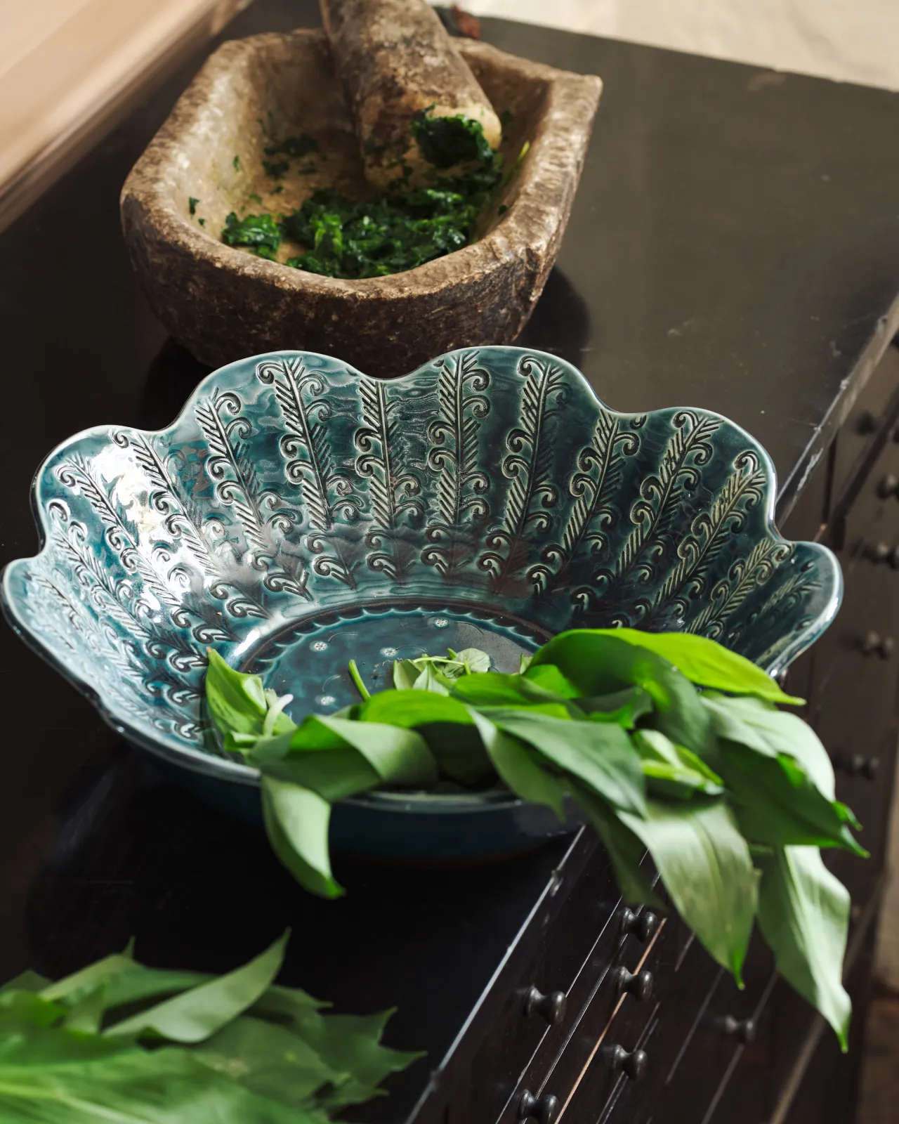 Blue glazed petal-shaped ceramic bowl with embossed pattern, styled with fresh wild garlic on a dark surface.