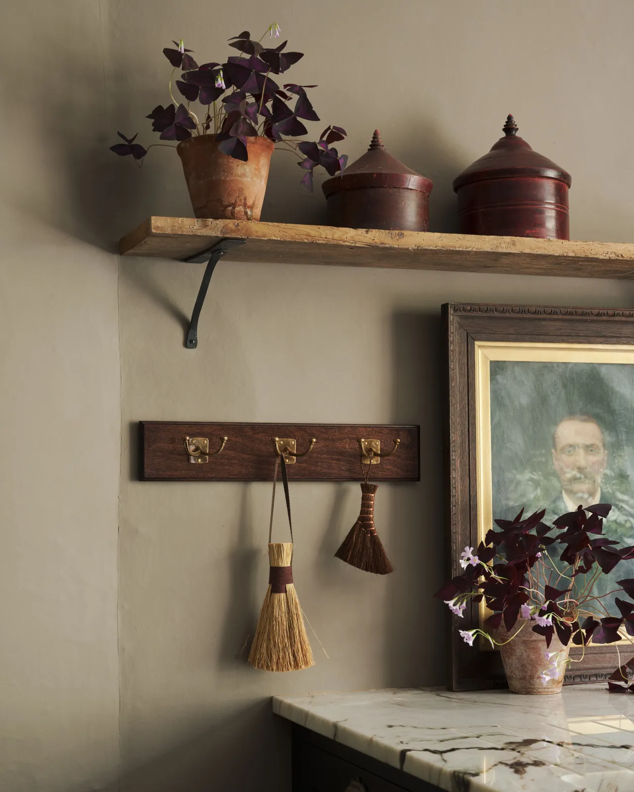 A Mahogany Finish backboard with three brass Twin Edwardian Hooks, fitted to a grey wall with a worn pine shelf, vintage potrait painting and potted plants.