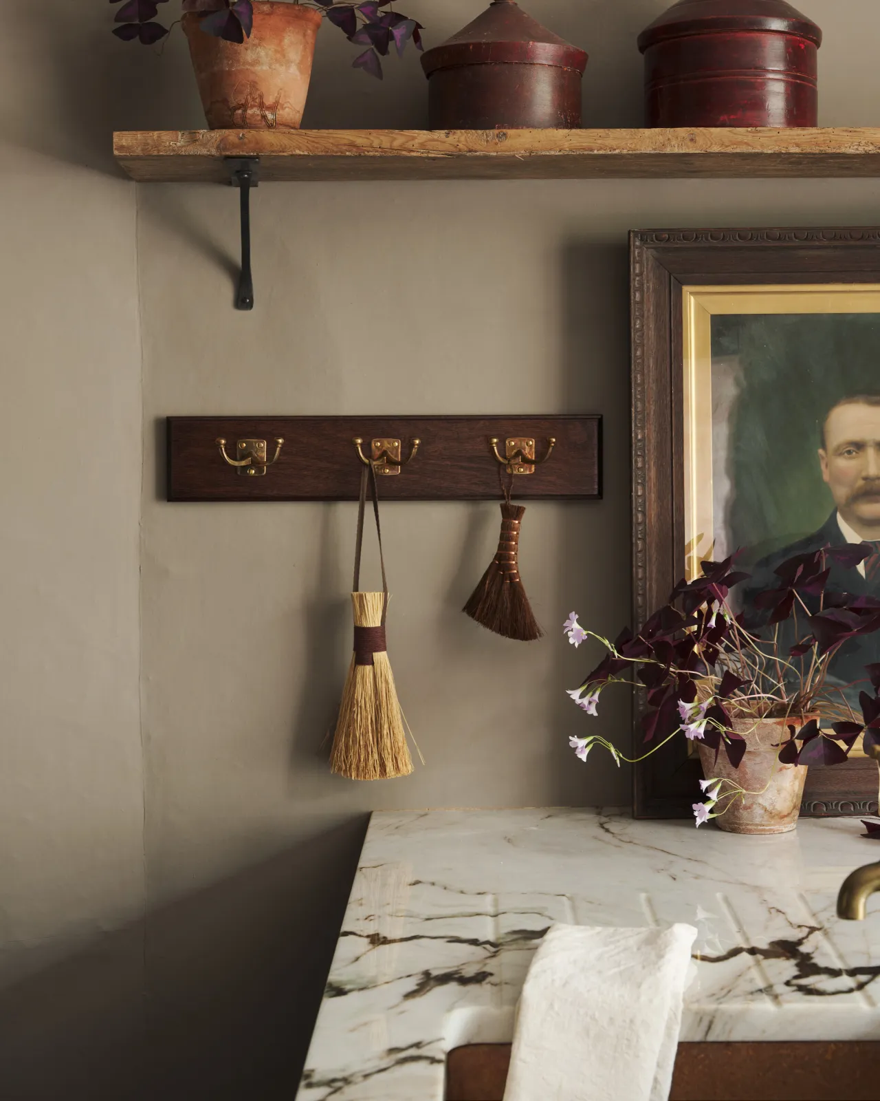 A Mahogany Finish backboard with three brass Twin Edwardian Hooks, fitted to a grey wall. Vintage brushes hang from the hooks, beside a small potted plant.