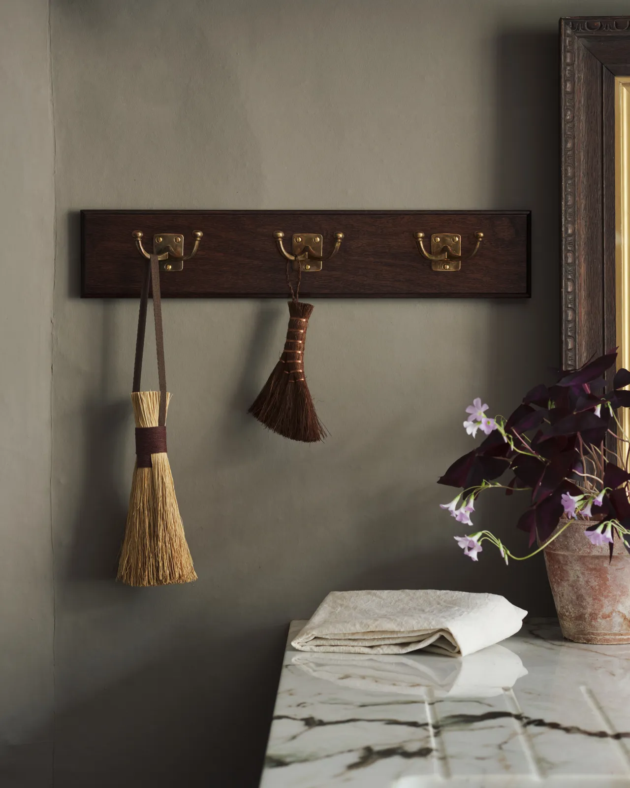 A close-up view of a Mahogany Finish backboard with three brass Twin Edwardian Hooks, fitted to a grey wall. Vintage brushes hang from the hooks, beside a small potted plant.