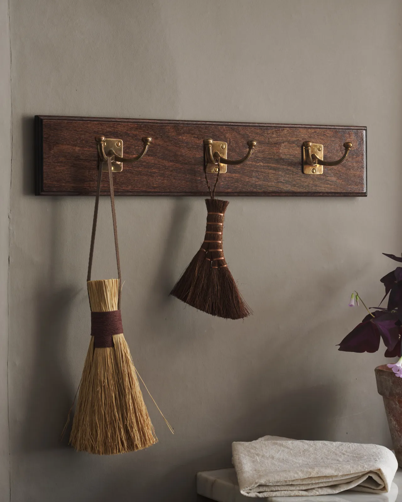 A close-up oblique view of a Mahogany Finish backboard with three brass Twin Edwardian Hooks, fitted to a grey wall. Vintage brushes hang from the hooks.