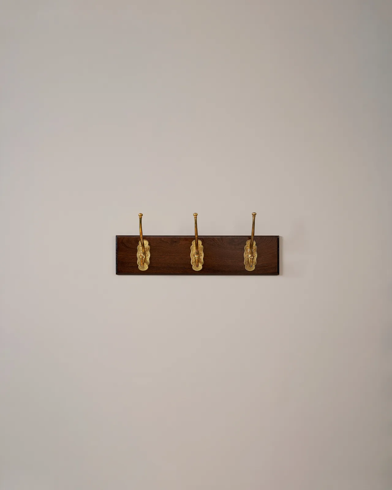 Three brass Grand Parisian Café Hooks on a Mahogany Finish Backboard on a plain grey wall.