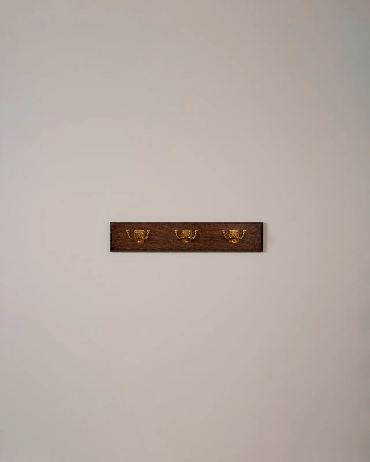 Three brass Twin Edwardian Hooks on a Mahogany Finish Backboard on a plain grey wall.