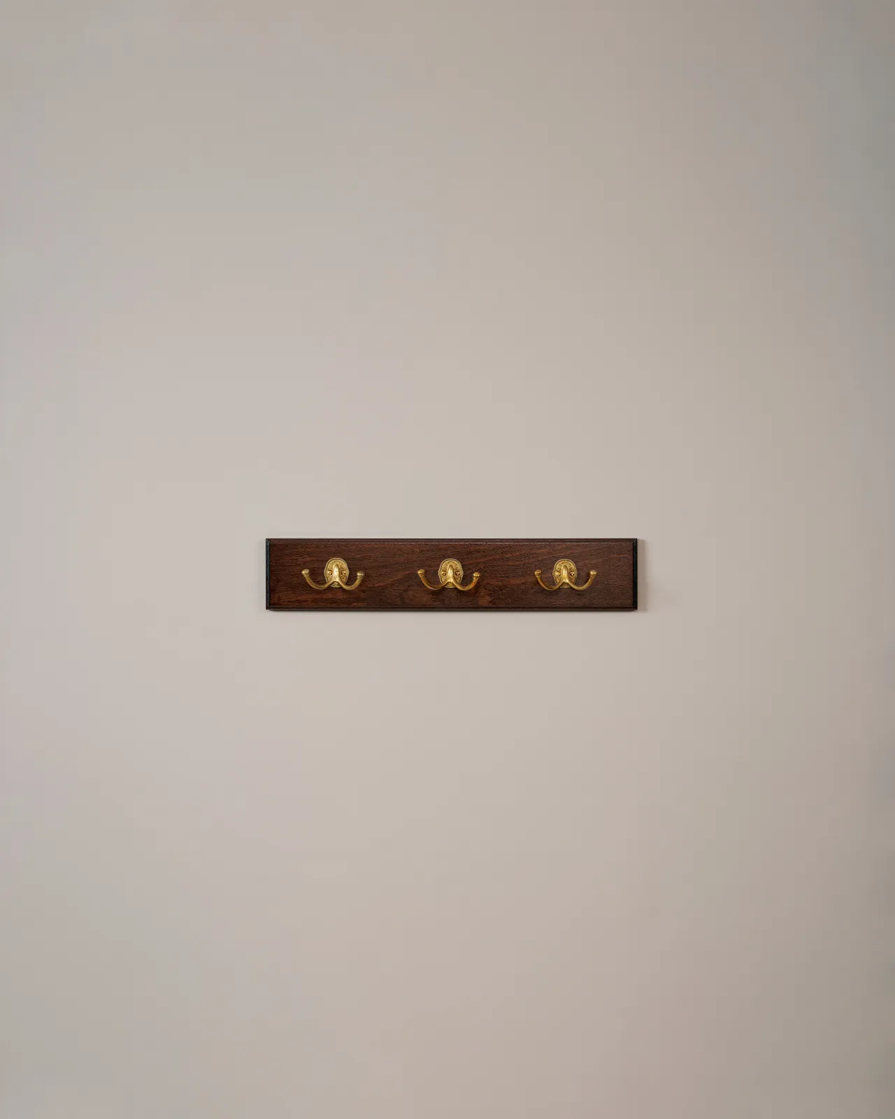 Three brass Twin Parisian Café Hooks on a Mahogany Finish Backboard on a plain grey wall.