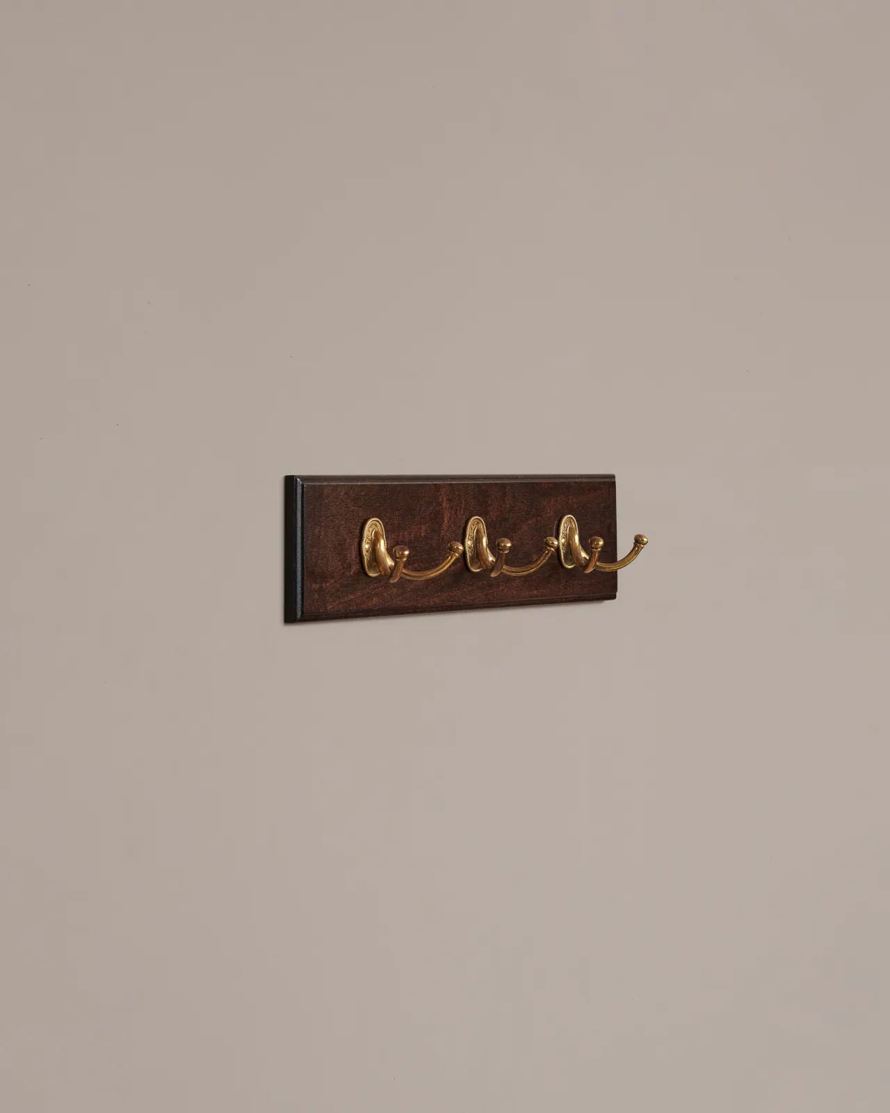 An oblique view of three brass Twin Parisian Café Hooks on a Mahogany Finish Backboard on a plain grey wall.
