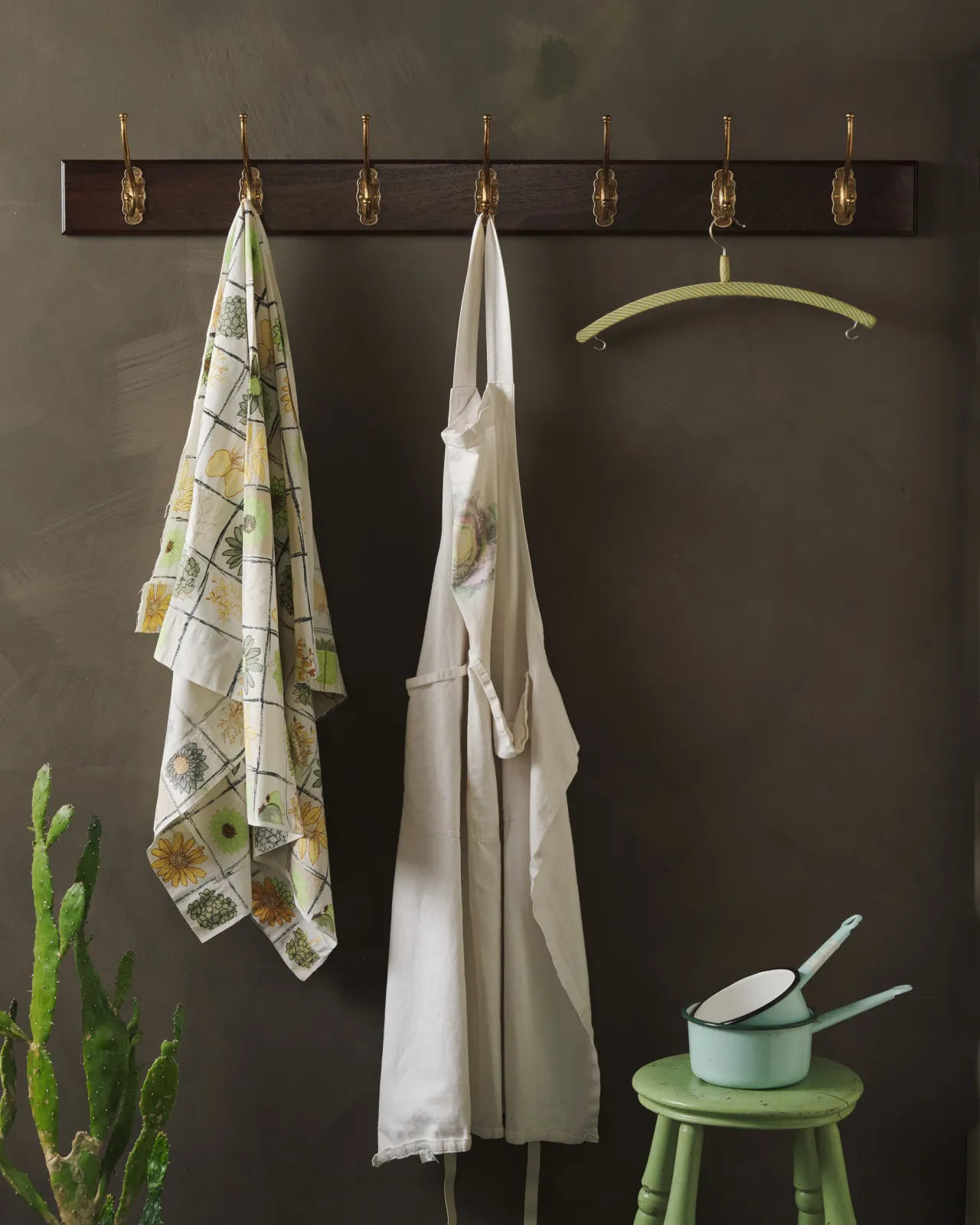 A Mahogany Finish Hook Rack with seven brass Parisian Café Hooks, fitted to a dark wall with two aprons hanging.
