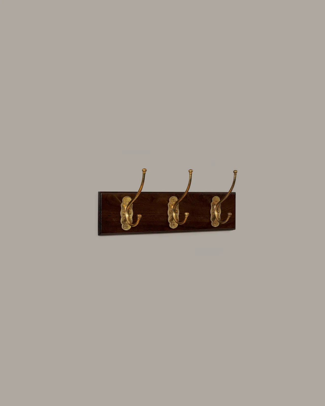 An oblique view of three brass Grand Parisian Coat Hooks on a Mahogany Finish Backboard on a plain grey wall.