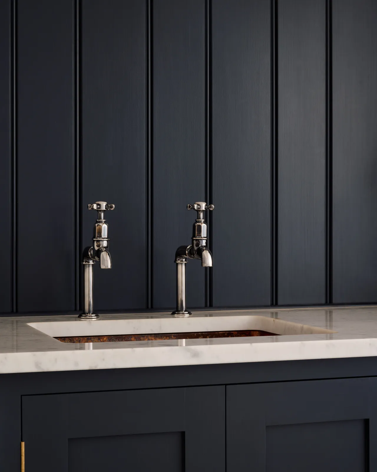 An oblique view of a pair of traditional-style tarnished silver bibcock taps with crosshead handles in a classic utility kitchen with a Carrara marble worktop and deep blue painted cupboards and tongue and groove panelling.
