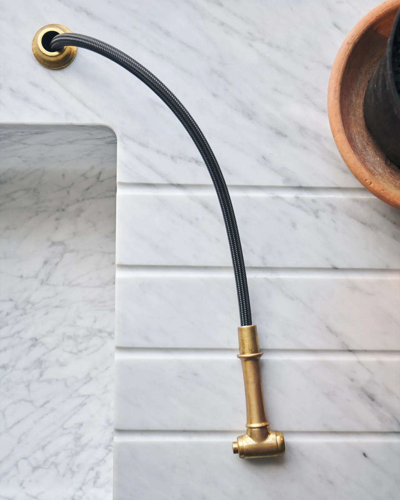 An overhead view of a traditional-style aged brass rinser tap and black hose, pulled out and resting on a Carrara marble worktop.