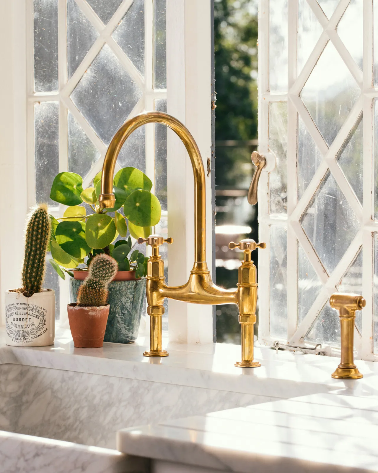 An oblique view of a traditional-style aged brass mixer tap with crosshead handles and an independent rinser tap on a bright Carrara marble worktop. Warm light pours in from an original Victorian leaded window behind the tap.