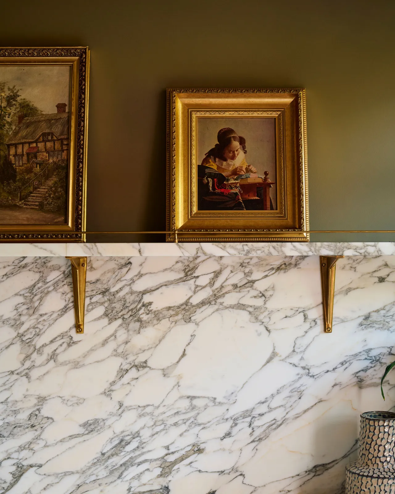 A straight-on view of an Arabescato marble splashblack and shelf with classic brass utilitarian shelf brackets and vintage oil paintings resting on top.