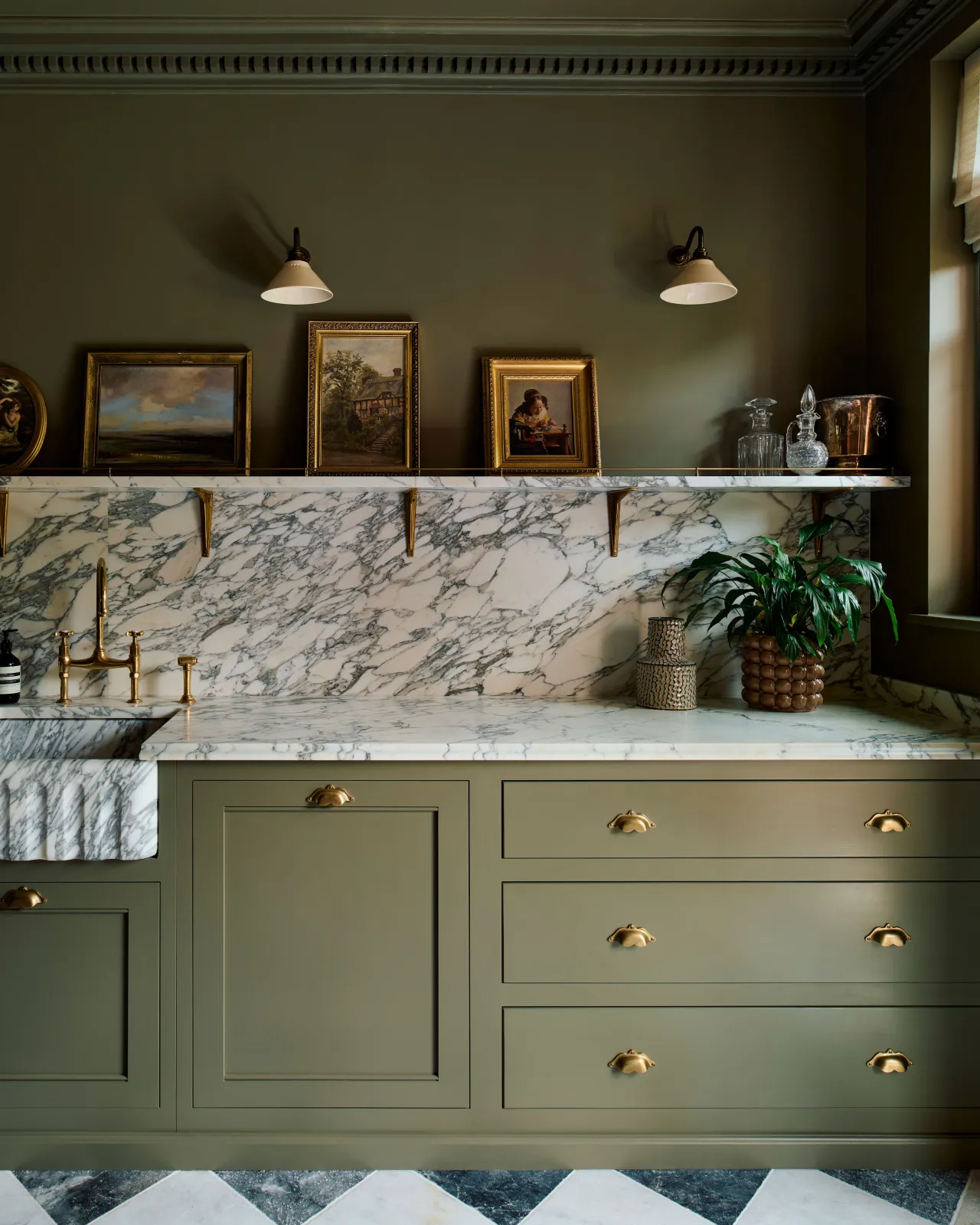 A straight-on view of a classic and moody Georgian-style kitchen with mossy green walls and cabinetry, an Arabescato marble worktop, splashback and shelf and classic brass utilitarian shelf brackets.